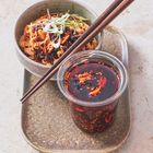 Eine Schüssel Nudeln mit schwarzer Soße, Frühlingszwiebeln. Daneben ein Glas mit roter Sojasoße. Dunkle Essstäbchen.