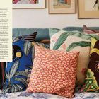 Der Text beschreibt die Vielseitigkeit und Dekorationsmöglichkeiten von Kissen auf einem Sofa. Verschiedene Kissen liegen bunt gemustert auf einem blau-grauen Sofa, das von gerahmten Kunstwerken überragt wird.