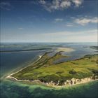 Keine erkennbaren Texte. Luftaufnahme einer grünen Insel mit Klippen und Wasser unter einem blauen Himmel mit einigen Wolken.