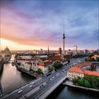 Aussicht auf eine Stadt mit einem großen Turm, Fluss, Brücke und historischen Gebäuden bei Sonnenuntergang.