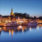 Abendstimmung mit beleuchteten Gebäuden, zwei weißen Booten im ruhigen Wasser und einer Kirche im Hintergrund.