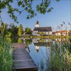 Ich kann keine Texte im Bild sehen. Eine Kirche am See mit einem Steg im Vordergrund, umgeben von Bäumen und Schilf.