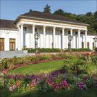 Text: "HAUS". Majestätisches Gebäude mit Säulen, umgeben von bunten Blumenbeeten und gepflegtem Rasen.