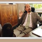 Ein Mann im Anzug sitzt entspannt in einem Büro, die Füße auf dem Schreibtisch, mit einem Aktenordner neben sich.