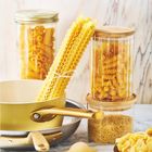 Verschiedene Sorten trockene Nudeln in Gläsern und auf Tellern, mit einer gelben Kasserolle und einer Pfanne im Hintergrund.