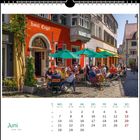 „Hotel Engel“, Juni 2021 Kalenderblatt, Menschen sitzen draußen, bunte Schirme, kopfsteingepflasterte Straße.