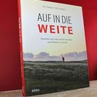 „AUF IN DIE WEITE“ in großen Buchstaben. Darunter: „Manchmal muss man einfach losziehen, um Antworten zu suchen.“ Landschaftsfoto mit Person.