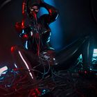 Eine Person in einem schwarzen Vinylanzug sitzt in einem Gewirr aus Kabeln, rot und blau beleuchtet.