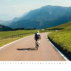 Text: "Juni", Zahlen 1-30 darunter. Ein Radfahrer auf einer kurvigen Straße durch grüne Hügel, Berge im Hintergrund. Logo rechts unten.