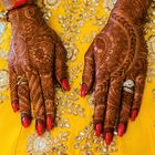 Zwei Hände mit Henna-Mustern und roten Nägeln ruhen auf goldbesticktem gelbem Stoff. Ringschmuck ist vorhanden.