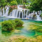 Keine Texte. Wasserfälle fließen über Felsen in smaragdgrünes Wasser, umgeben von dichtem, grünem Laub.