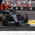 Text: "JUNI" und Kalendertage darunter. Ein Formel-1-Auto mit der Nummer 63 fährt vor Streckenposten in orangefarbenen Overalls.