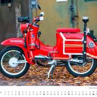 „FEUERWEHR“ steht auf einem roten Moped geparkt auf Laub. Unten ist ein Kalender für Dezember.