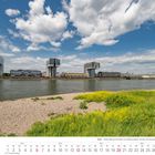 Text: Juni 2027, Köln - Poller Wiesen mit Blick auf die Kranhäuser.  
Vordergrund: Wiese und Kies, Hintergrund: Rhein, moderne Architektur.