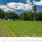 6 2027. Kalenderblatt. Ein Feld mit grünen Pflanzen, ein Baumreihe und ein Wasserturm bei blauem Himmel mit Wolken.