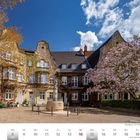 März 2027 Kalender. Pauliplatz mit Panbrunnen in Köln-Braunsfeld. Gepflasterter Platz, blühender Baum, historische Gebäude.