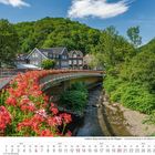 Kalenderblatt für Juni 2027, Bild zeigt eine Brücke mit roten Blumen, einen Fluss und Häuser vor bewaldeten Hügeln.