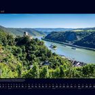 07 Juli. Rheinlandschaft mit Burg und Fluss. Weinberge im Vordergrund, Berge im Hintergrund, Boote auf dem Wasser.