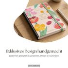 „Exklusives Design handgemacht“, Notizbuch mit bunten Blumen auf einem Holztisch, daneben ein Stift.