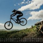 "FOX" auf Kleidung, "STREET" verschwommen auf Holz. Kalender unten. Radfahrer springt in grüner Landschaft vor blauem Himmel.