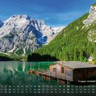 Text: "Pragser Wildsee, Dolomiten". Holzhütte am türkisgrünen See, umgeben von Bergen und Wäldern, mit Ruderbooten am Steg.