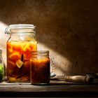 Drei Einmachgläser auf Holztisch; links grün, Mitte orange mit Vanilleschote, rechts rot. Halbschatten.