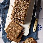 Ein Brotlaib mit Sonnenblumenkernen darauf, teilweise geschnitten. Daneben ein Messer auf blauer Serviette. Fliesenhintergrund.