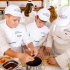 "Drei Kinder in weißen Kochjacken mit Namen Benedikt, Lorenz, Mia. 'Europa Miniköche' auf Kappen und Jacken. Schneiden Tomaten."