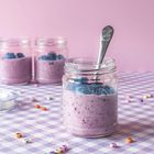 Buchstabenperlen auf lila-weiß karierter Decke, Glas mit lila Joghurt, Heidelbeeren und Löffel, rosa Hintergrund.