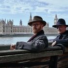 Zwei Männer mit Hüten sitzen auf einer Bank am Fluss. Im Hintergrund die Häuser des Parlaments in London.