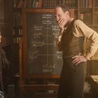 Tafel mit Notizen, Mann in nachdenklicher Pose, Frau daneben, Bücherregale im Hintergrund, warme Beleuchtung.