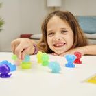 Ein lachendes Mädchen spielt mit bunten Plastikfiguren auf einem Tisch. Im Hintergrund sind ein Sofa und Pflanzen zu sehen.