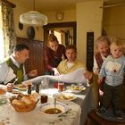 Eine Familie sitzt um einen Tisch mit Essen, während eine Person ein großes Papier betrachtet. Ein Kleinkind steht auf einem Stuhl.