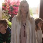 Eine Frau mit langen, blonden Haaren steht neben zwei Kindern in einem Raum mit großen Fenstern und Blick auf bunte Blumen.