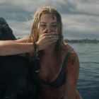 Eine Frau sitzt erschöpft an einem Felsen im Meer, bedeckt mit Wunden, den Mund mit der Hand haltend. Wolkiger Himmel.