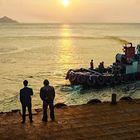 Zwei Personen stehen auf einem Pier und blicken auf ein Boot und den Sonnenuntergang über dem Meer.