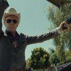 Ein Mann mit Cowboyhut und Sonnenbrille hält eine Hand hoch und eine Pistole in der anderen. Im Hintergrund Bäume.