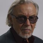 Eine ältere Person mit grauen Haaren und Sonnenbrille, in einem dunklen Anzug mit rotem Hemd.