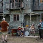 Drei Männer hören einer Band mit Gitarren und Schlagzeug zu, die vor einem alten, verwitterten Haus im Freien spielt.