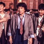 Fünf Männer in Anzügen stehen unsicher und besorgt eng beieinander, während Gewehre auf sie gerichtet sind.