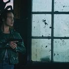 Eine Frau mit grüner Lederjacke hält eine Pistole, steht neben einem schmutzigen Fenster in dunklem Raum.
