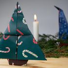 Origami-Tannenbaum aus grünem Papier mit Zuckerstangenmuster, Tannenzweige und eine brennende Kerze im Hintergrund.