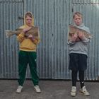 Zwei Jungen stehen vor einer Wellblechwand, jeder hält einen großen Fisch. Einer trägt einen gelben Hoodie, der andere grau.