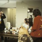 Ein Mann steht gestikulierend, trägt ein "Spinal Tap" T-Shirt. Andere sitzen um einen Tisch mit Essen und Getränken.