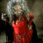 Ein fiktives Wesen mit langem, zerzaustem Haar, trägt ein rotes Gewand und hat ein ungewöhnliches, detailreiches Gesicht.