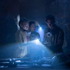 Drei Personen mit Taschenlampen und Atemmasken untersuchen ein Skelett in schwach beleuchtetem, mysteriösem Raum.