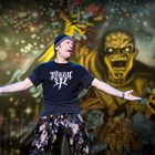 T-Shirt text: "Bite the Remains". Mann mit Mikrofon, bunter Bühnenhintergrund mit Monster-Illustration im Stil eines Gemäldes.