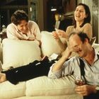 Ich kann den Text nicht erkennen. Drei Menschen lachen auf einem weißen Sofa; einer hält ein Getränk, andere einen Zettel.