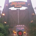 Schriftzug "JURASSIC PARK" über einem großen, hölzernen Tor im Regen. Ein Geländewagen fährt hindurch, umgeben von Palmen.