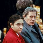 Zwei Personen sitzen auf Stühlen, umgeben von herbstlichem Laub. Eine junge Frau in Rot, ein älterer Mann mit grauem Mantel.